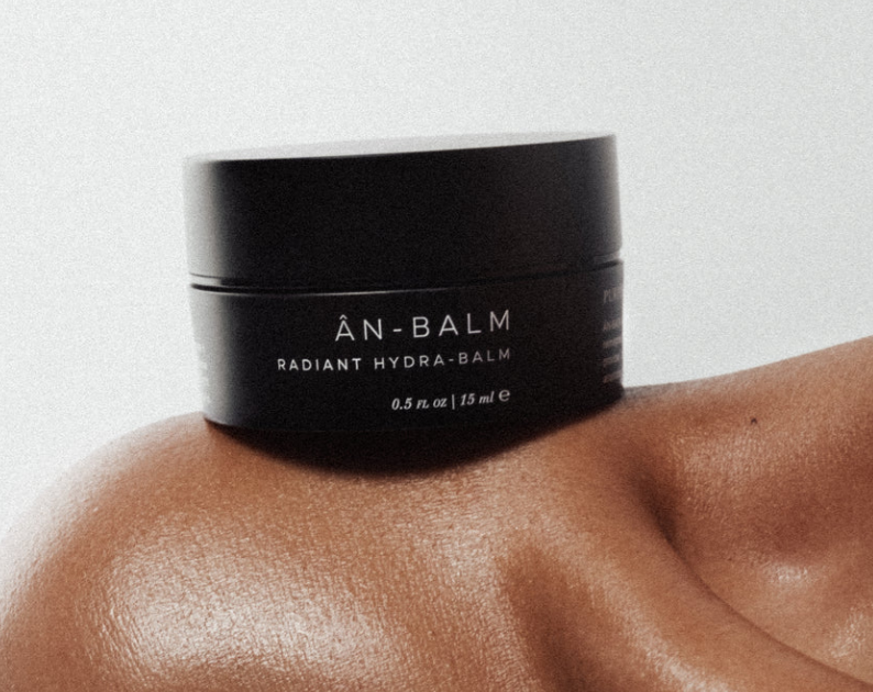 Black jar of 'AN-BALM Radiant Hydra-Balm' on a person's hand against a light background